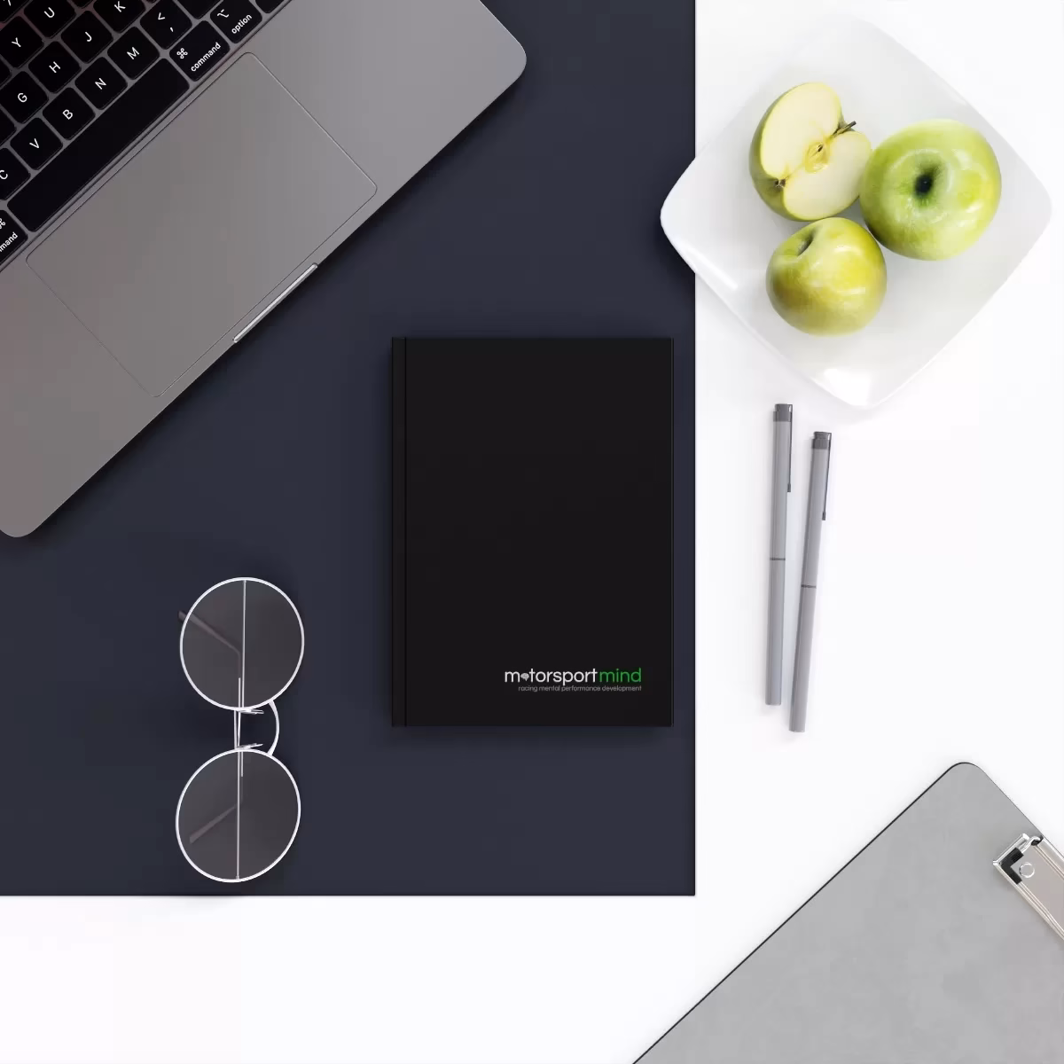 A hard backed journal on a desk next to an apple and a laptop.